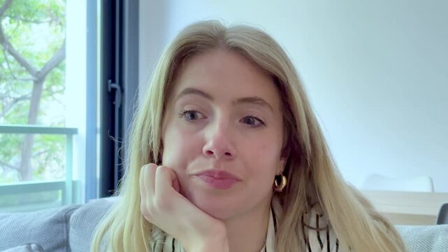 Frustrated blonde woman sitting on the couch in her living room, displaying boredom and impatience while waiting for something, with apathetic and tired expressions on her face