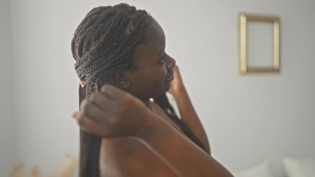 Woman with long braided hair touches braid and smiles, bare shoulders visible in a building; confidence natural selfcare.