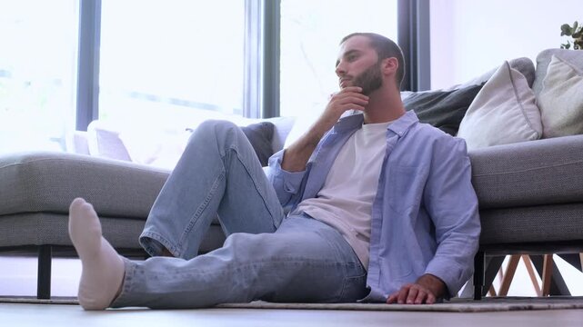 Stressed young man sitting on the living room floor, feeling overwhelmed and anxious while experiencing intense neck pain and discomfort from mental health issues and overthinking