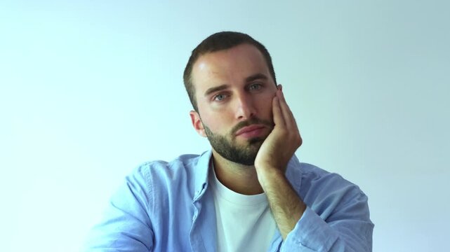 Unhappy caucasian man with a beard sitting at a table isolated on a white background, looking at the camera with a sad and pensive expression, feeling bored, lonely and depressed