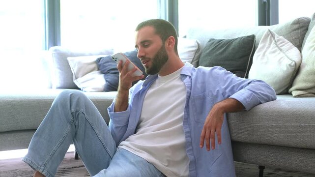 Handsome caucasian man relaxing in his modern apartment, sitting on the floor leaning against the sofa while talking on speakerphone or recording a voice message on his mobile phone