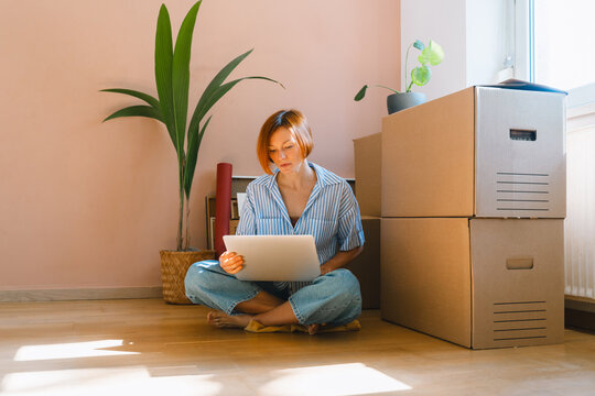 A young woman sits on the wooden floor using her laptop surrounded by packed cardboard boxes. Authentic first home ownership and real estate property investment concept.