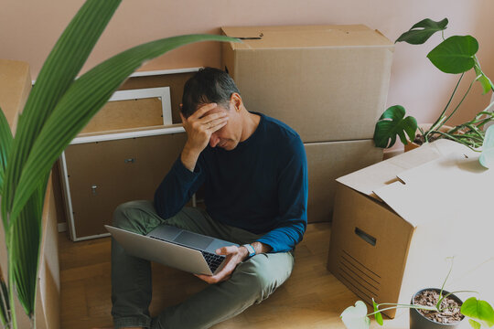 A frustrated man holds his head while sitting on the floor with a laptop surrounded by boxes. Authentic home relocation and real estate mortgage pressure concept.