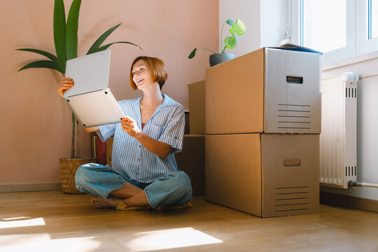 A young woman sits on the wooden floor using her laptop surrounded by packed cardboard boxes. Authentic first home ownership and real estate property investment concept.
