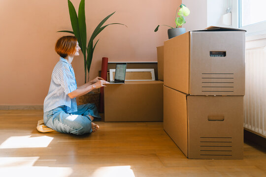 A young woman sits on the wooden floor using her laptop surrounded by packed cardboard boxes. Authentic first home ownership and real estate property investment concept.