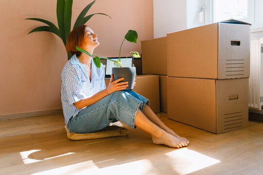 A happy young woman holds a small potted plant while sitting on the floor among cardboard boxes. Authentic first home ownership and real estate property concept.