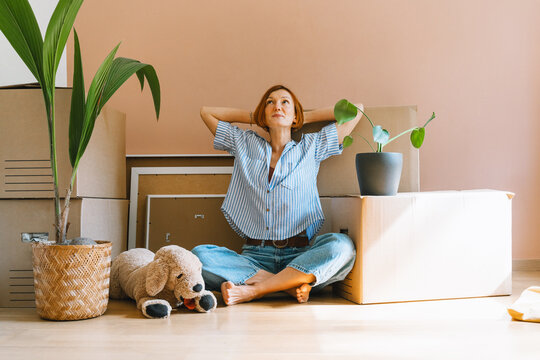 A happy young woman sits on the floor with plush dog toy next to a potted houseplant. Authentic first home ownership and real estate property investment concept.