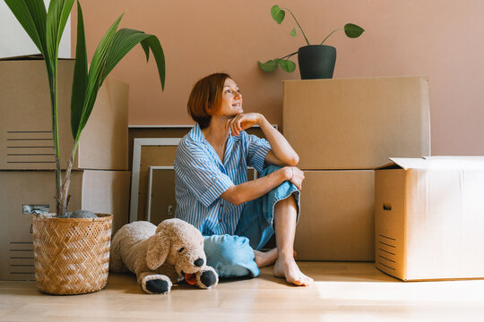 A happy young woman sits on the floor with plush dog toy next to a potted houseplant. Authentic first home ownership and real estate property investment concept.
