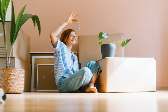 A joyful young woman raises her new house keys high while sitting on the floor among boxes. Authentic first home ownership and real estate property investment concept.