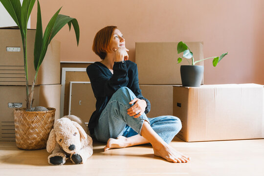A thoughtful young woman in a black shirt sits on the floor looking up among moving boxes. Authentic first home ownership and real estate property investment concept.