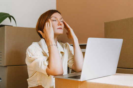 A stressed young woman rubs her head while looking at a laptop among packed cardboard boxes. Authentic home relocation and real estate mortgage pressure concept.