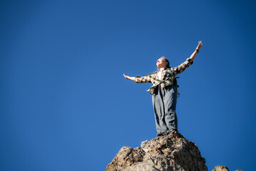 Obraz premium Successful female traveler celebrates reaching a rocky mountain summit with arms raised toward the sky. Adventure travel, personal achievement, freedom, confidence and outdoor exploration concept.