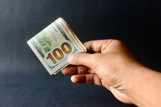 Hand offering a folded stack of 100 US dollar bills on a dark background.