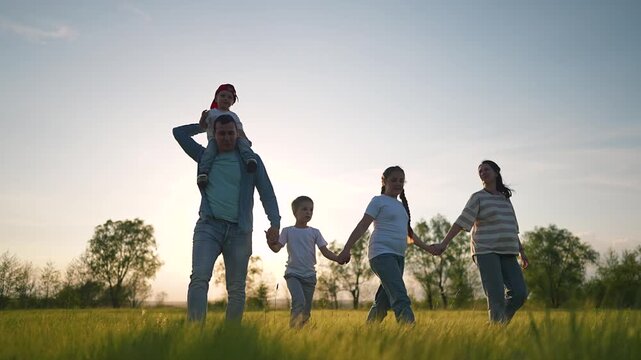 Mother walking through green meadow holding hands family. Sunset lights calm evening walk. Family moves slowly through tall grass. Mother holds daughter and son while walking across peaceful field.