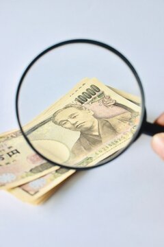 A magnifying glass inspecting a Japanese 10000 yen banknote close-up, visualizing financial scrutiny, foreign currency evaluation, and economic analysis.
