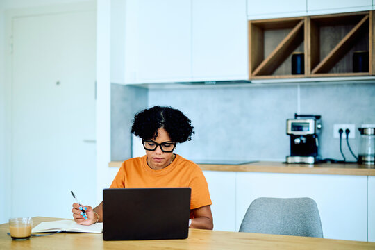 Woman concentrating on her laptop and writing notes in a notebook, working from her home kitchen with a coffee