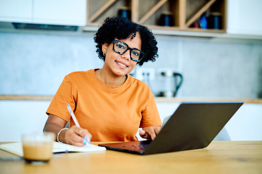 Young woman with glasses smiling at camera, writing notes in a notebook while working on a laptop at her home office