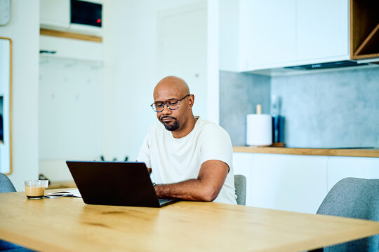 Adult man with glasses concentrating on his laptop, working remotely from a home office setup in a bright kitchen