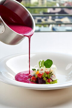 Pouring bright pink beetroot cream soup into a white bowl garnished with fresh greens and microgreens. High-end gourmet dining.