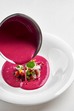 Pouring bright pink beetroot cream soup into a white bowl garnished with fresh greens and microgreens. High-end gourmet dining.