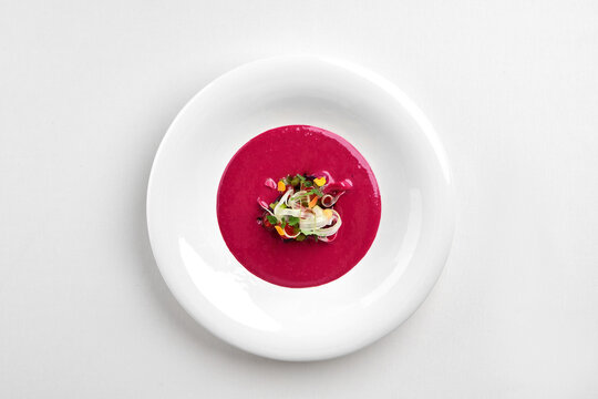 Gourmet pink beetroot cream soup served in a white bowl, garnished with microgreens and fresh herbs. Isolated on white background.