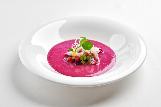 Gourmet pink beetroot cream soup served in a white bowl, garnished with microgreens and fresh herbs. Isolated on white background.