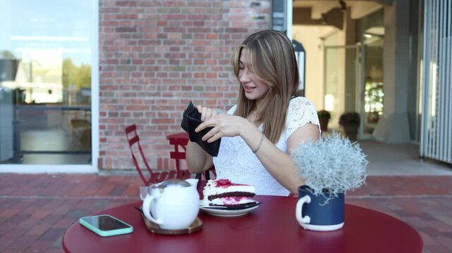 Young woman checks her empty wallet at outdoor cafe after ordering cake and coffee looking worried she has no money to pay the bill and facing an awkward financial problem
