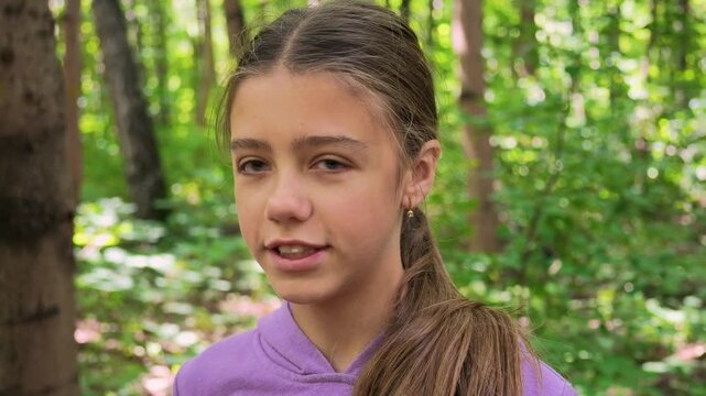 Teenage girl in purple hoodie talking while walking through a sunlit green forest, turning from camera toward side profile during an outdoor nature walk