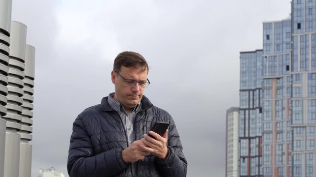 Middle aged man wearing glasses using smartphone outside and celebrating success with fist pump gesture