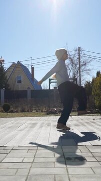 Cheerful young girl in knit hat jumping, dancing, and playing on sunny day in paved backyard