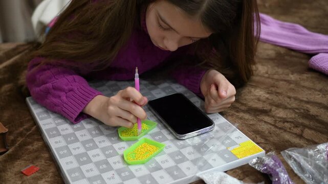 Young girl lying on bed at home, carefully applying small yellow rhinestones to diamond painting