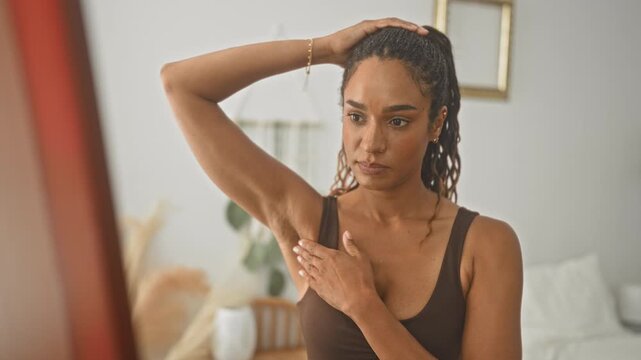 Woman touching armpit and examining skin in front of mirror in studio with natural light and plant; self care concern routine.