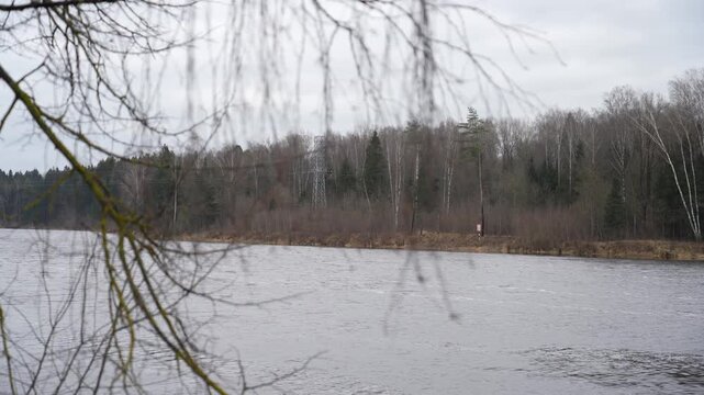 Wide river flowing slowly past bare autumn forest and power line tower on cloudy overcast day