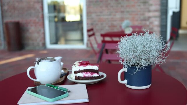 Empty cafe table with raspberry chocolate cake teapot smartphone notebook and pen beside a decorative plant creating a quiet workspace for a relaxing afternoon tea breaks