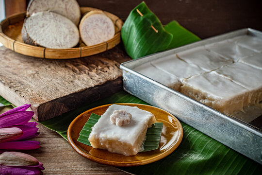 Traditional Thai tako taro pudding dessert square cut topped with thick coconut milk cream in aluminum tray next to banana leaf parcels and fresh sliced taro root. Asian sweet food snack.