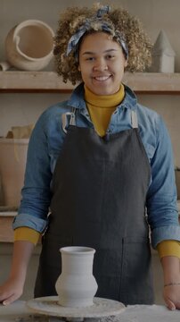 Slow motion portrait of cheerful African American woman in apron standing in pottery workshop next to throwing wheel smiling looking at camera