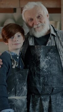 Slow motion portrait of senior man and child wearing aprons standing in pottery workshop smiling looking at camera. Family business and culture concept.