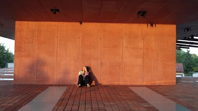 Woman sits against a wall in warm sunset light holding a smartphone and touching her head while reacting with stress and frustration to troubling news on the screen today