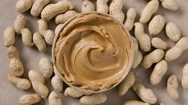 Smooth creamy peanut butter in a rotating bowl with a swirl on top, peanuts in the background. top view