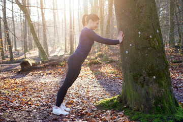 Fototapeta premium Athletic young sporty woman doing stretching exercises on a tree in forest on a sunny winter morning