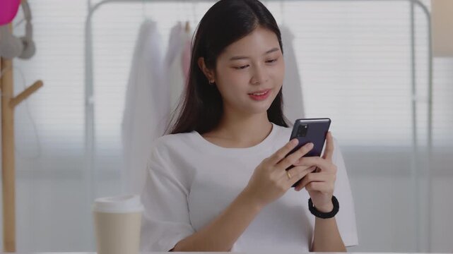 Smiling young Asian woman using smartphone to interact with followers during beauty live stream or social media management.