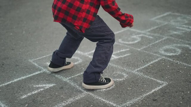 Child playing hopscotch on street. Kid steps on chalk hopscotch grid with black shoes. Outdoor activity for children, playing hopscotch in the sun. Fun and active hopscotch game for kids on the street