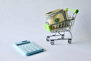 Mini shopping cart filled with US cash placed near a light blue digital calculator, symbolizing retail expense calculation, budgeting, and shopping costs. © Akram