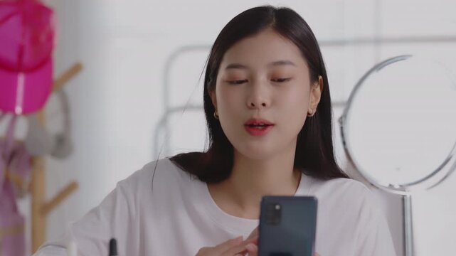 Young Asian beauty vlogger applying blush with a makeup brush while recording tutorial on smartphone.