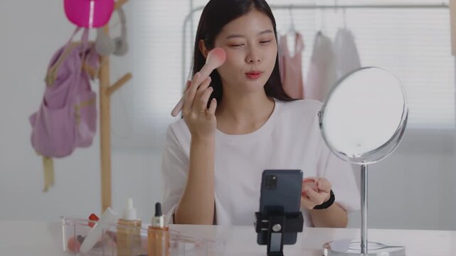 Young Asian beauty vlogger applying blush with a makeup brush while recording tutorial on smartphone.
