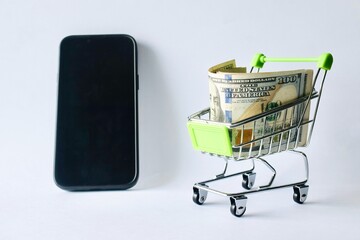 Black smartphone standing upright beside a miniature shopping cart loaded with US dollar banknotes on a white surface. © Akram