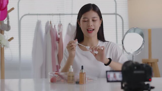 Young Asian beauty vlogger presenting cosmetic product in front of a digital camera for live streaming or video recording.