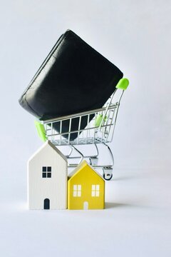Mini miniature house models in front of a shopping cart carrying a black leather wallet, representing real estate market trends, home buying, and personal mortgage budget management.
