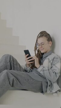 Joyful young woman with dreadlocks using smartphone checking online content sitting on stairs in luxury house. Modern technology and youth concept.