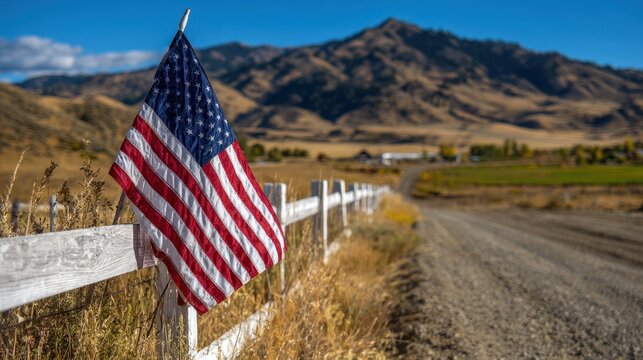 American flag on a white wooden fence along a rural dirt road with scenic mountains and blue sky, capturing patriotic country life and western landscape beauty.